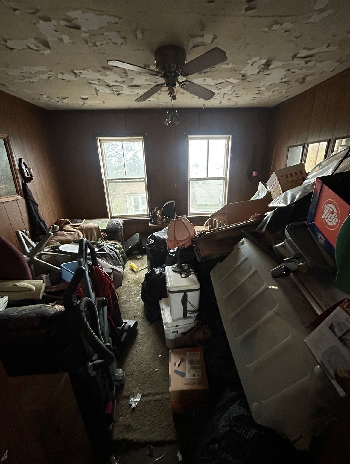 Cluttered room with peeling ceiling, two windows, and piles of items blocking the view.