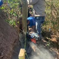 Two men are working on a wooden post in the dirt.