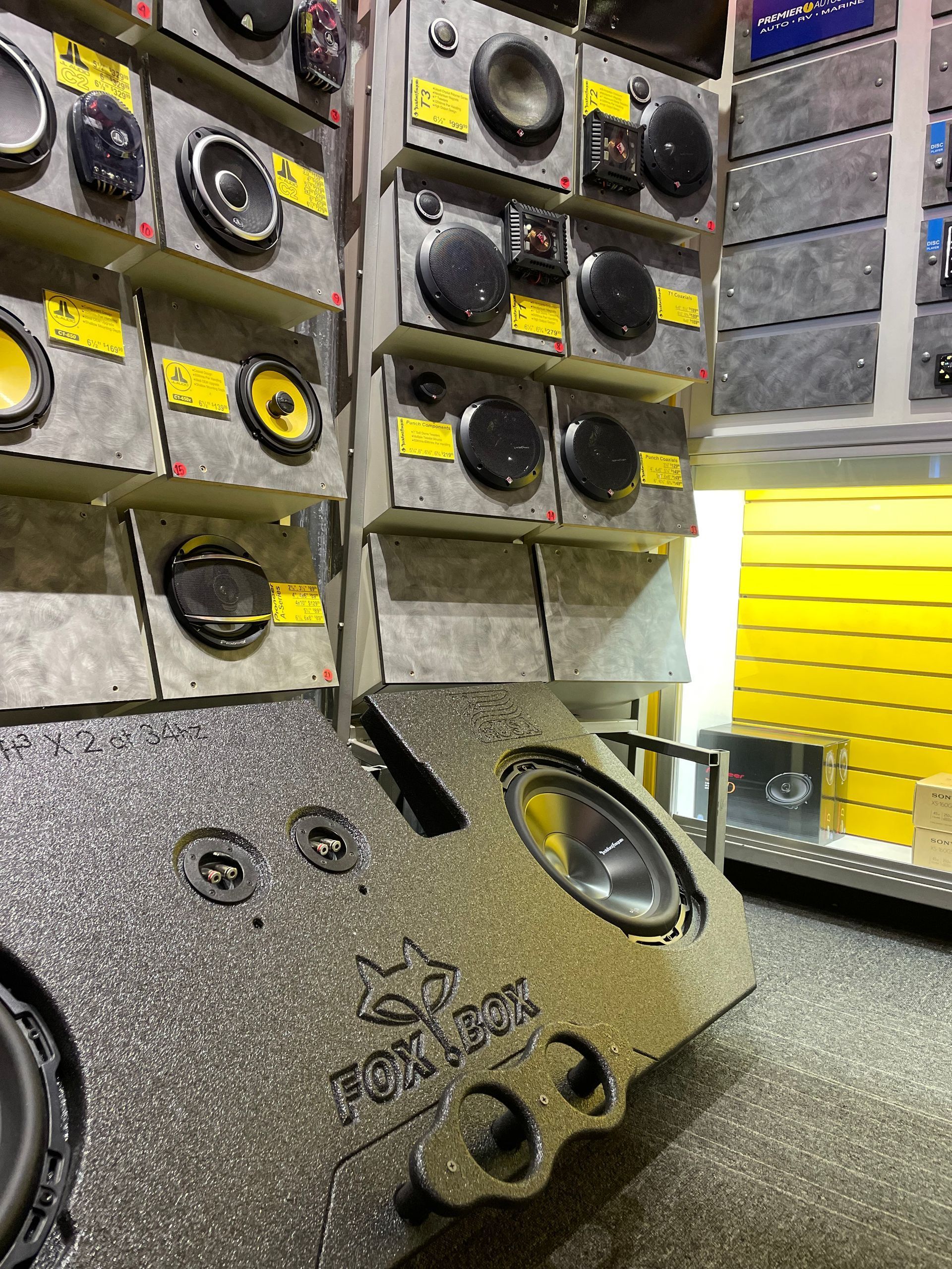 A display wall in an electronics store featuring various car audio speakers, subwoofers, and a branded bass enclosure.