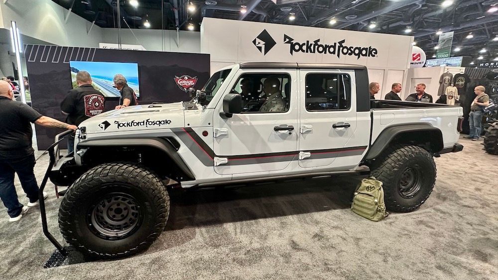 A white Jeep Gladiator pickup truck with large off-road tires and custom decals on display at a trade show booth.