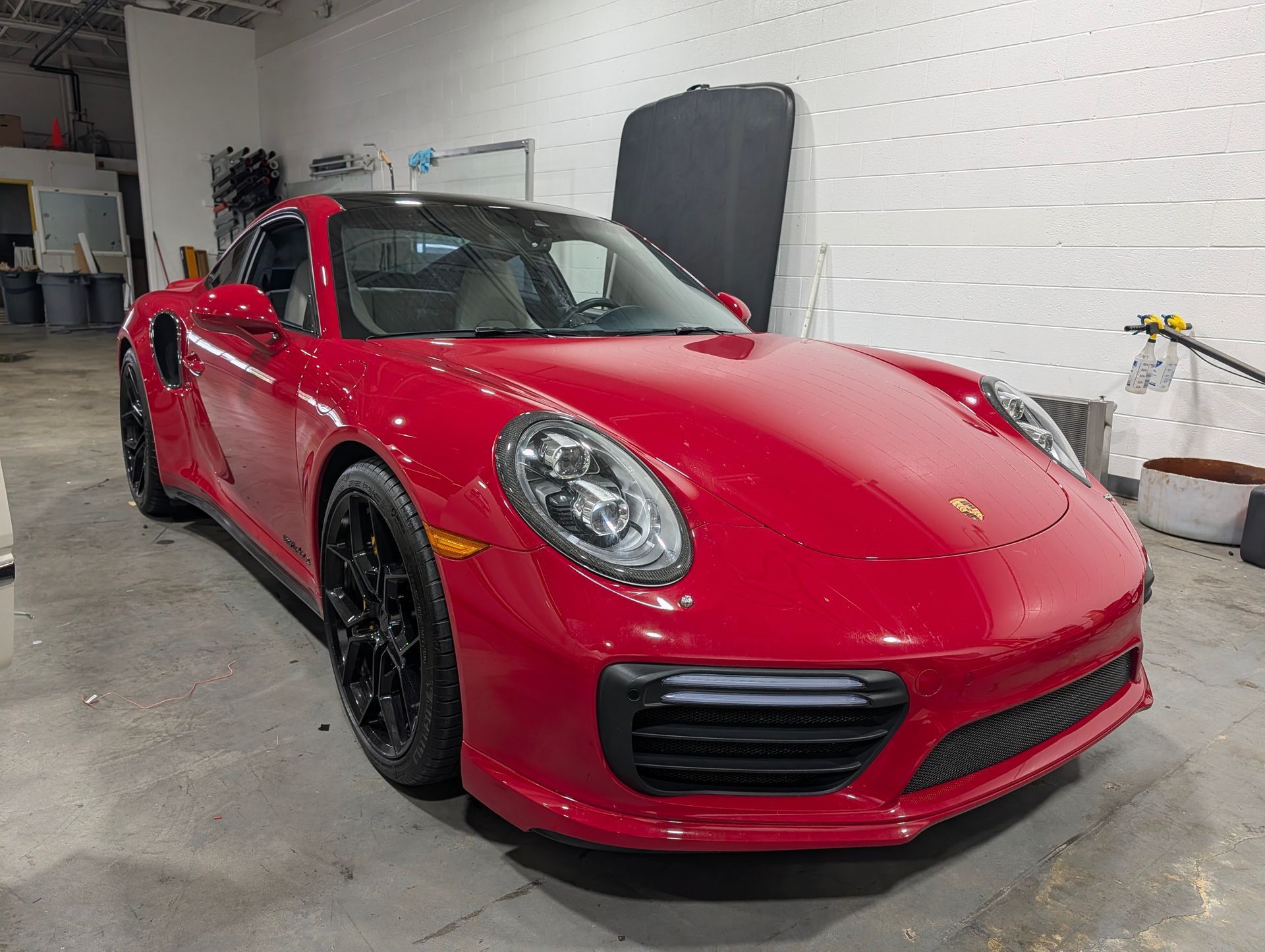 Red sports car in a garage, front-left view, with black wheels and a white brick wall background