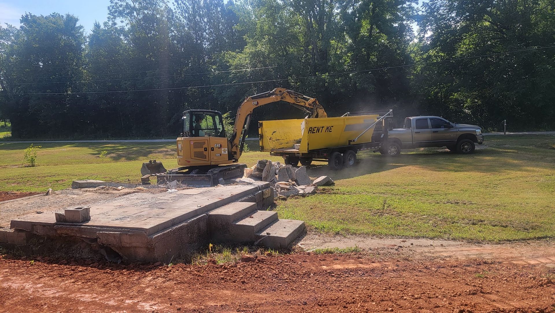 Excavator and yellow dump truck at a construction site beside a road, with stacked concrete slabs in front.