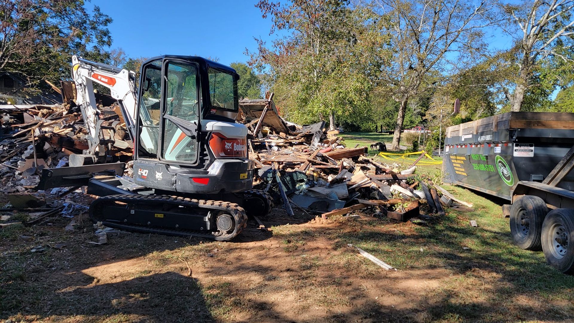 Mini excavator clearing debris beside a dump trailer in a wooded cleanup site