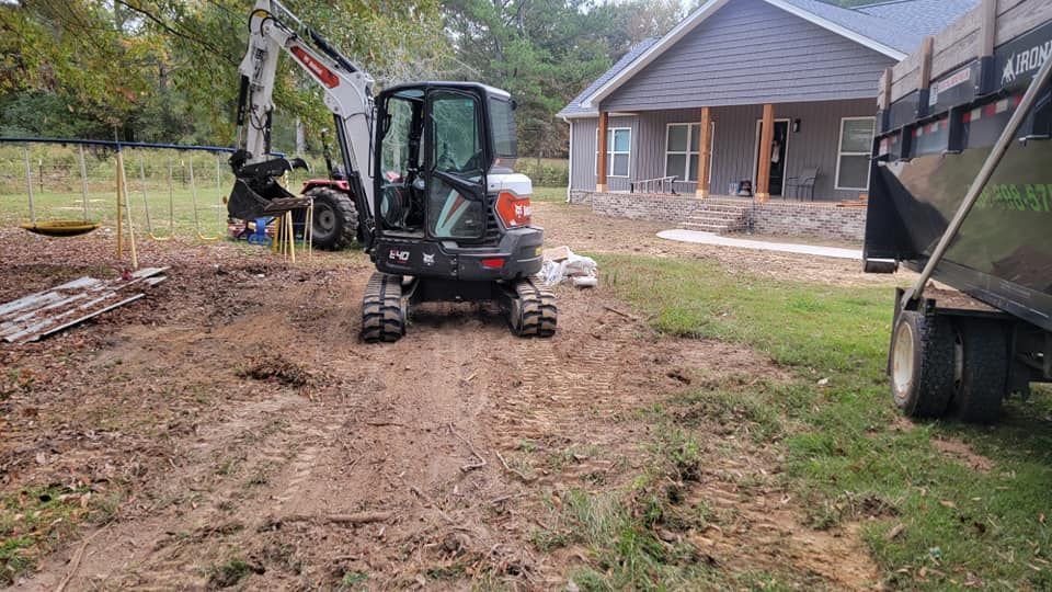 Mini excavator on muddy driveway beside a house, with a dump trailer on the right.