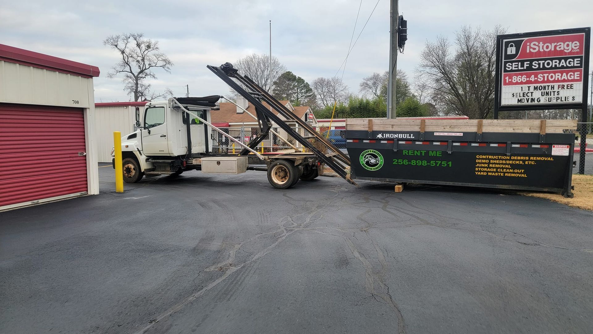 Dumpster truck unloading debris beside a building, with a green dumpster and wet parking lot.