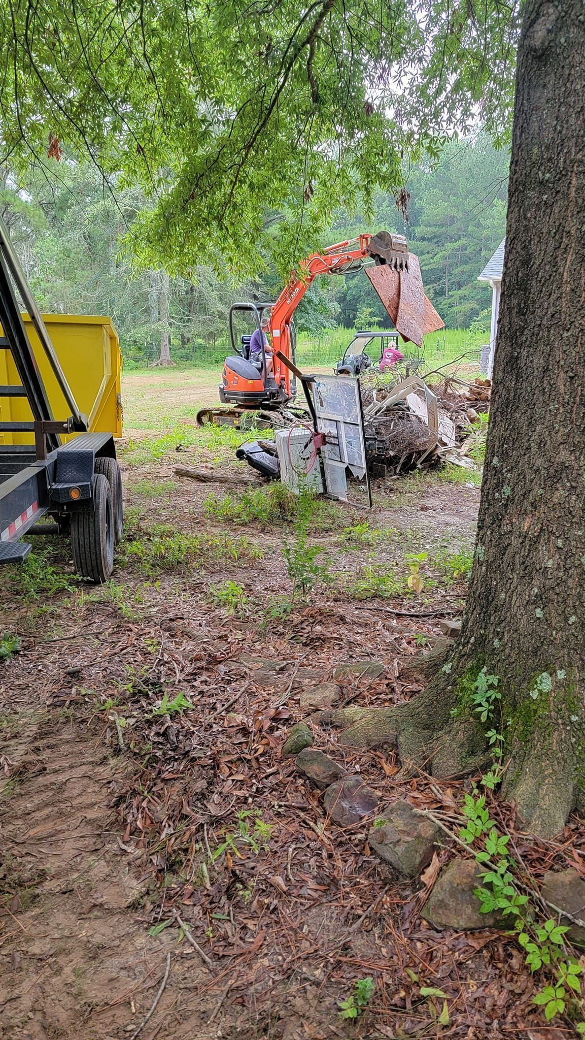 Orange excavator clearing brush in a wooded area, with a yellow vehicle partly visible at left.