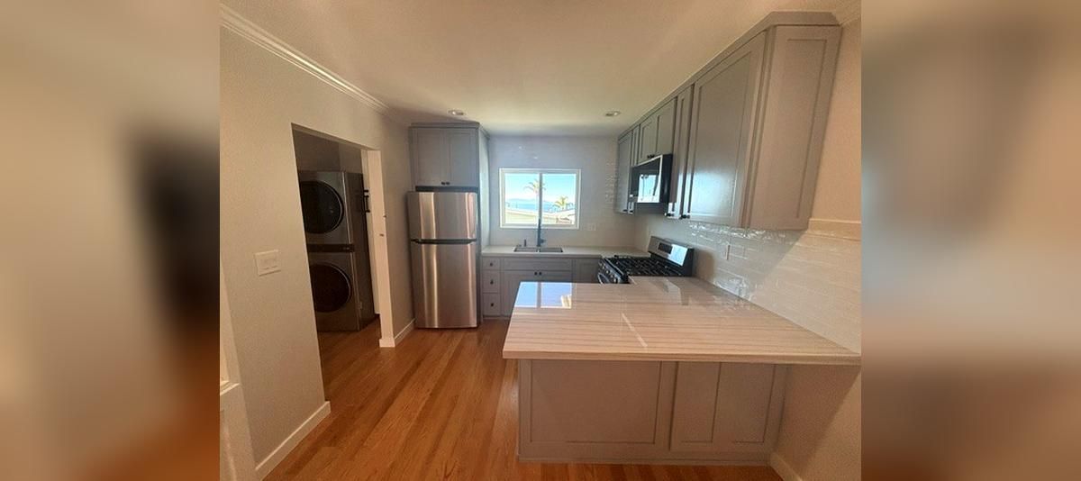 Bright kitchen with white cabinets, stainless fridge, and wood floors, viewed toward a windowed sink area.