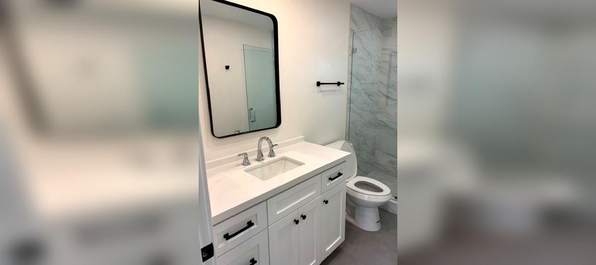 Bright bathroom with white vanity, mirror, sink, and toilet beside gray tiled walls.