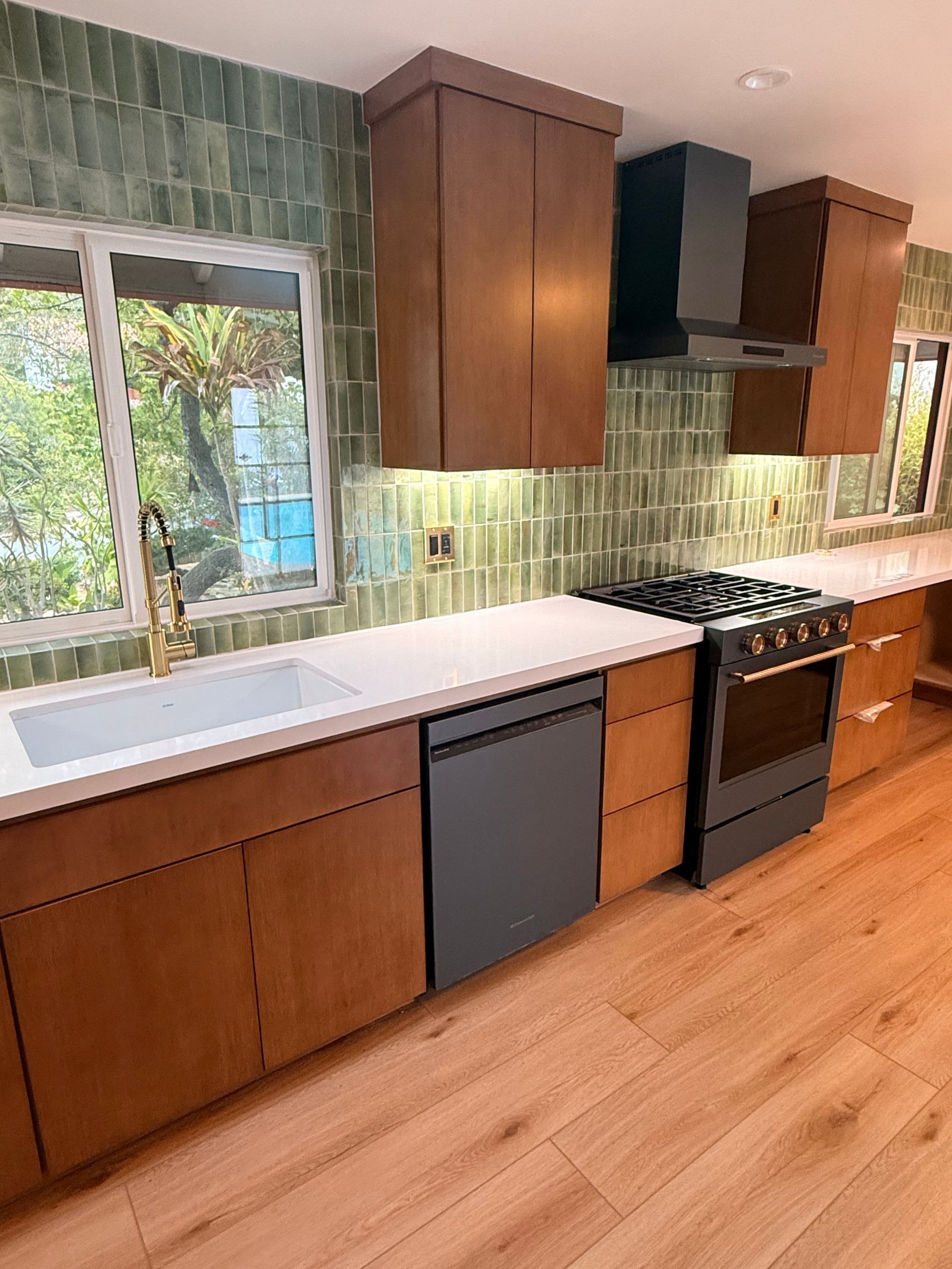 Modern kitchen with wood cabinets, green tile backsplash, black stove, and large window above a white countertop.