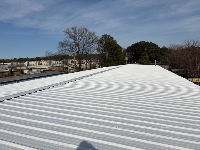 A bright white, reflective metal roof on a building, seen from a high angle under a blue, sunny sky with trees in the back.