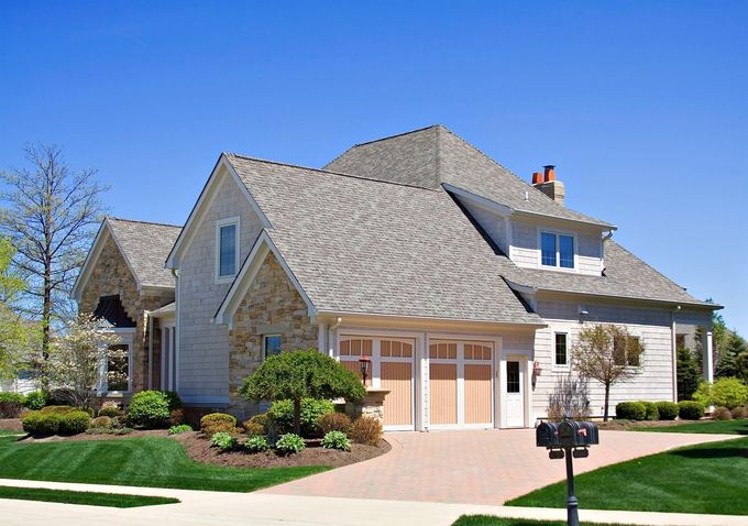 A two-story house with stone and light-colored siding, a shingled roof, a two-car garage, and a manicured front lawn.
