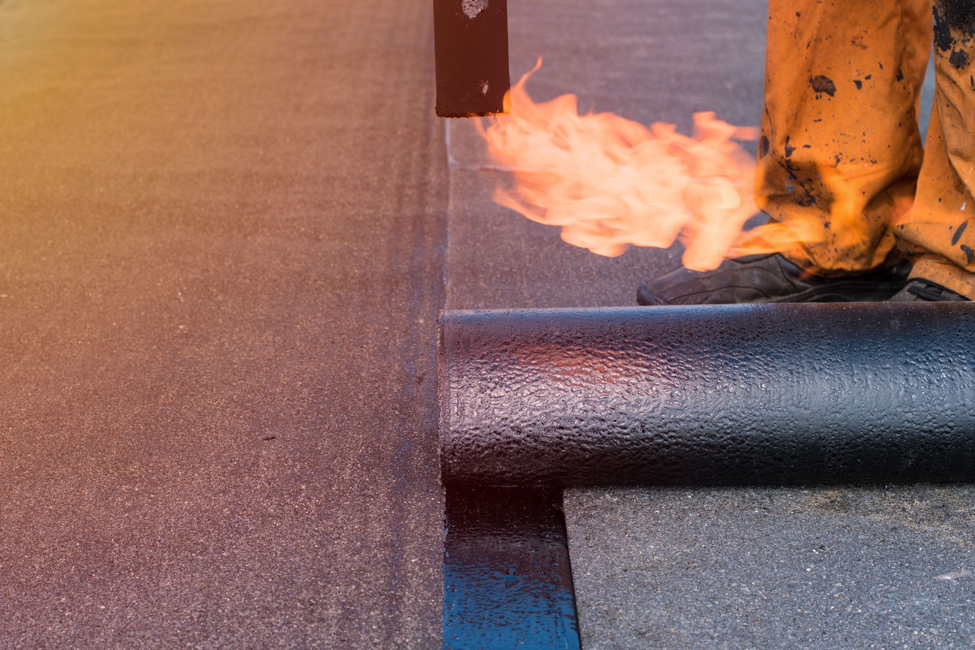 Roofer using a torch to install roofing material on a dark surface; fire flares near the rolled material.