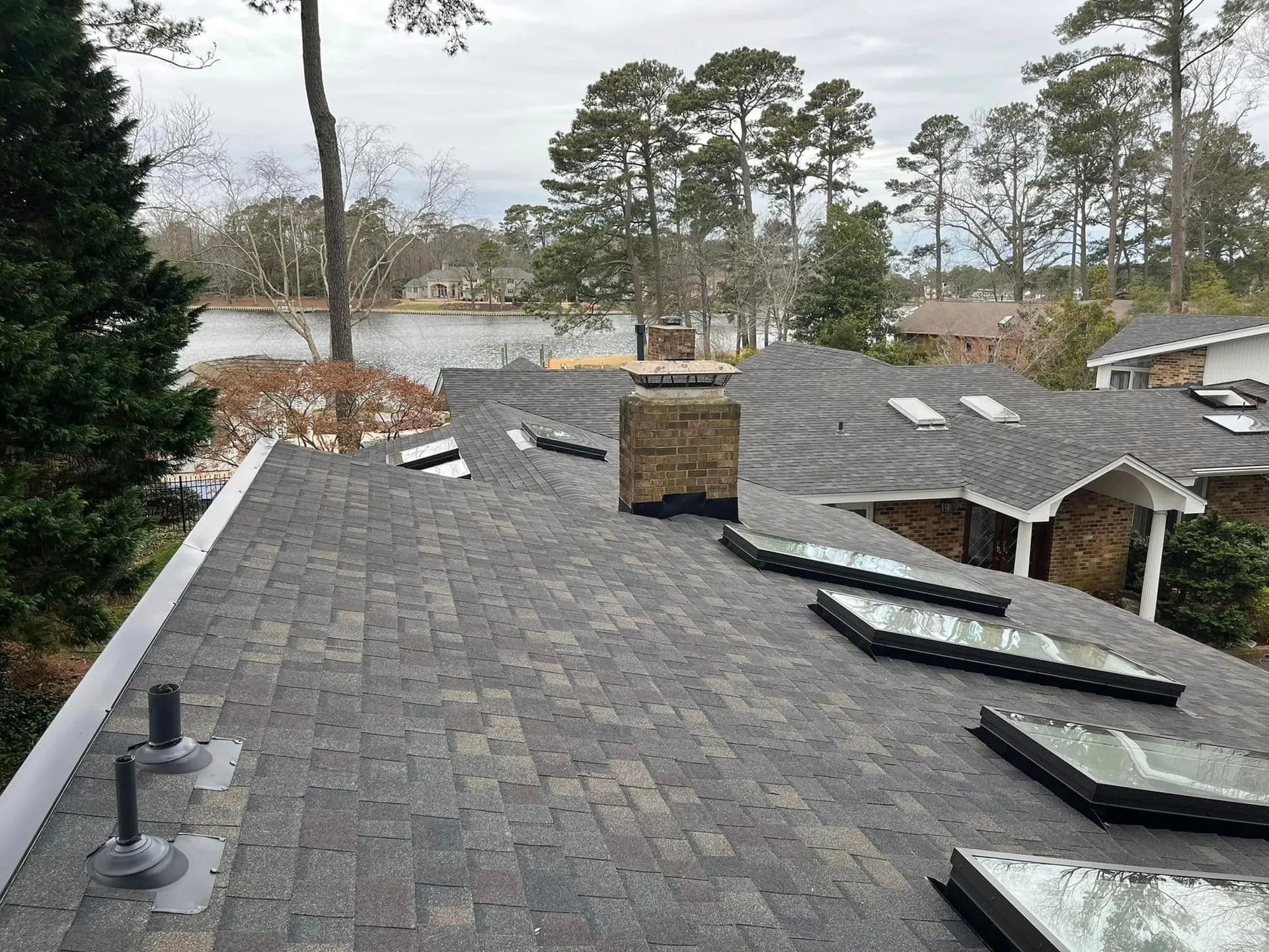 Rooftop view with dark shingles, brick chimney, skylights, and a lake in the background.