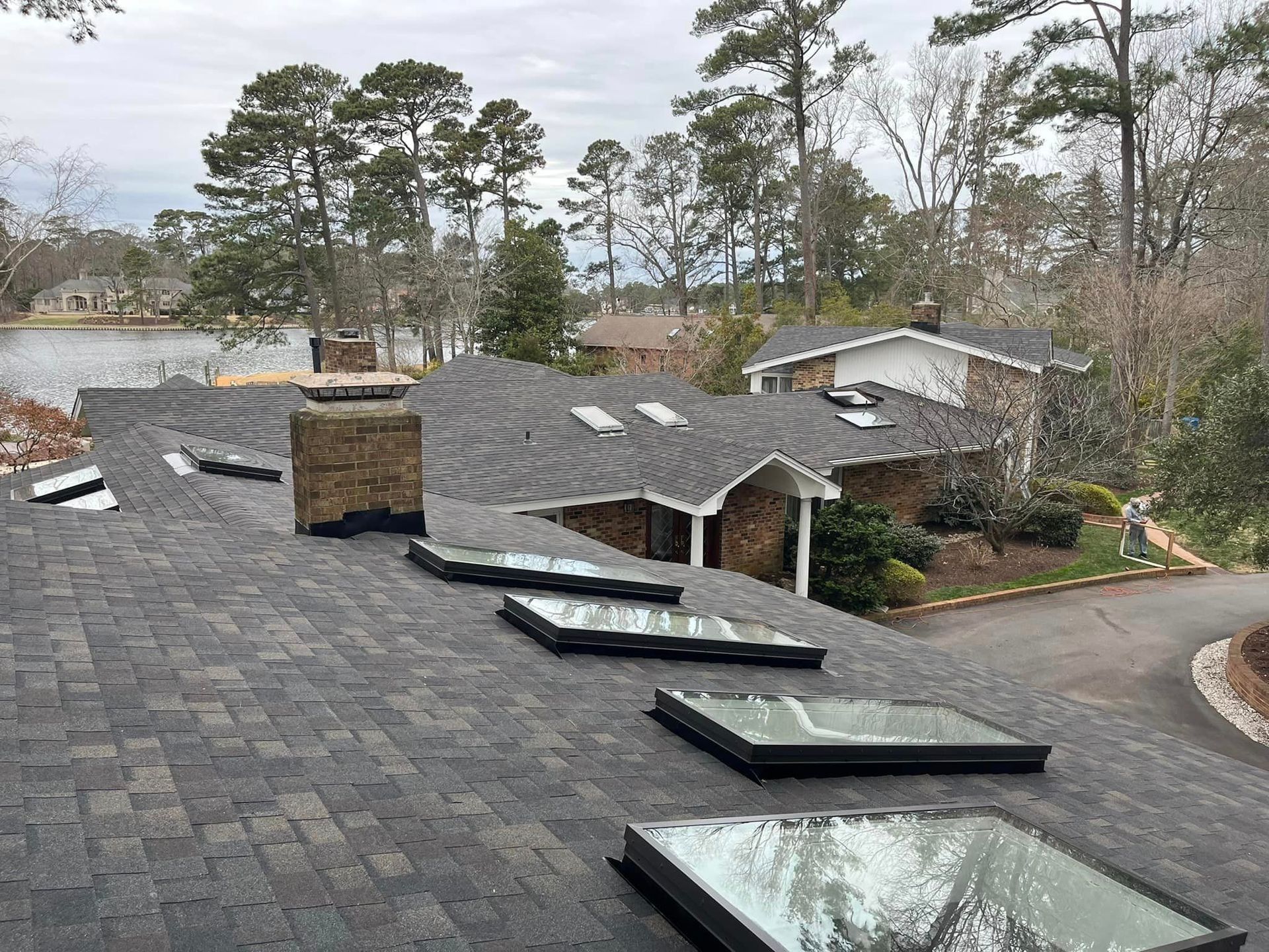 Gray shingled roof with skylights and brick chimneys, overlooking a lake with trees.