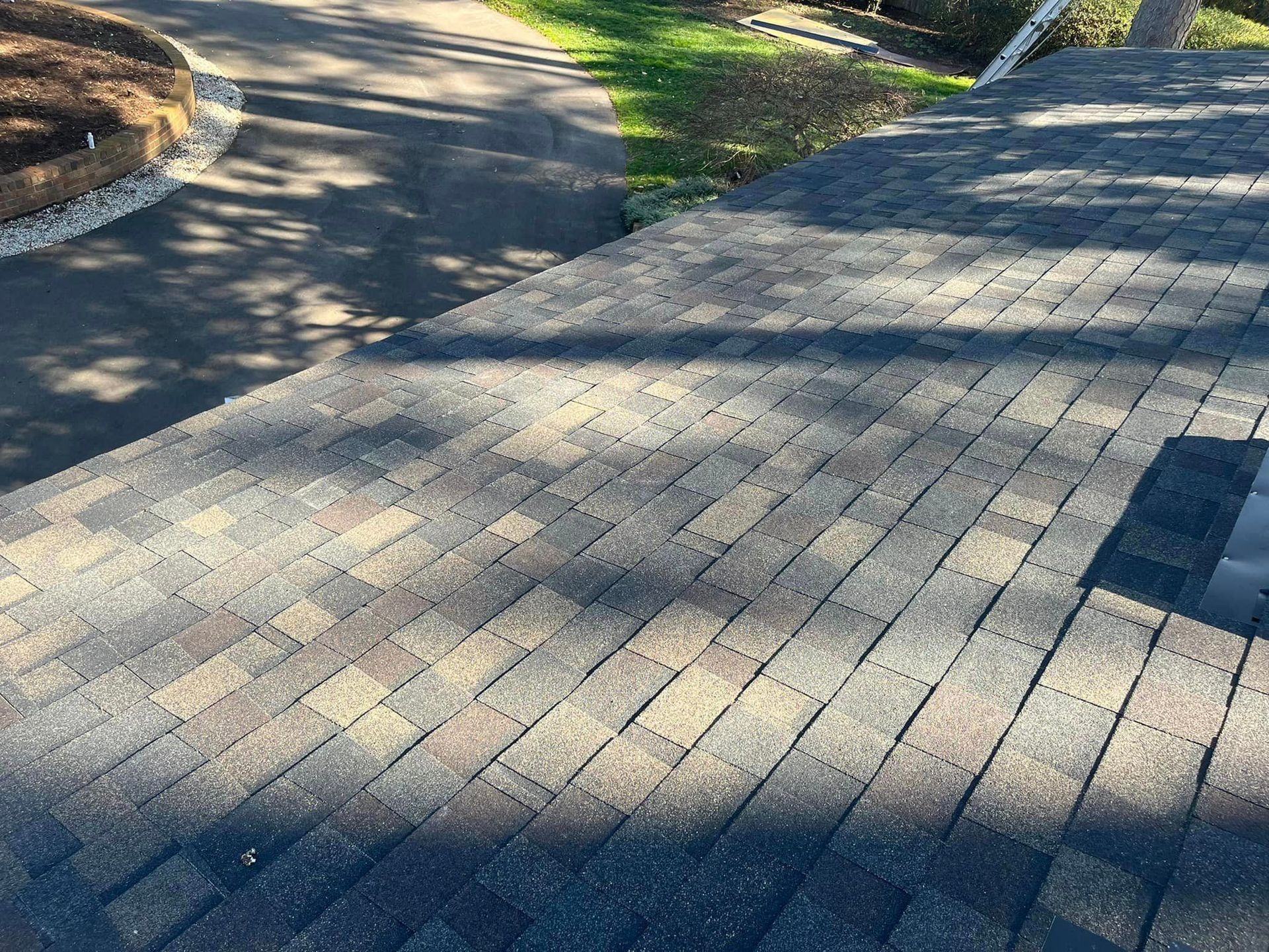 Dark asphalt shingle roof with varying shades of brown and gray; a driveway and yard in the background.