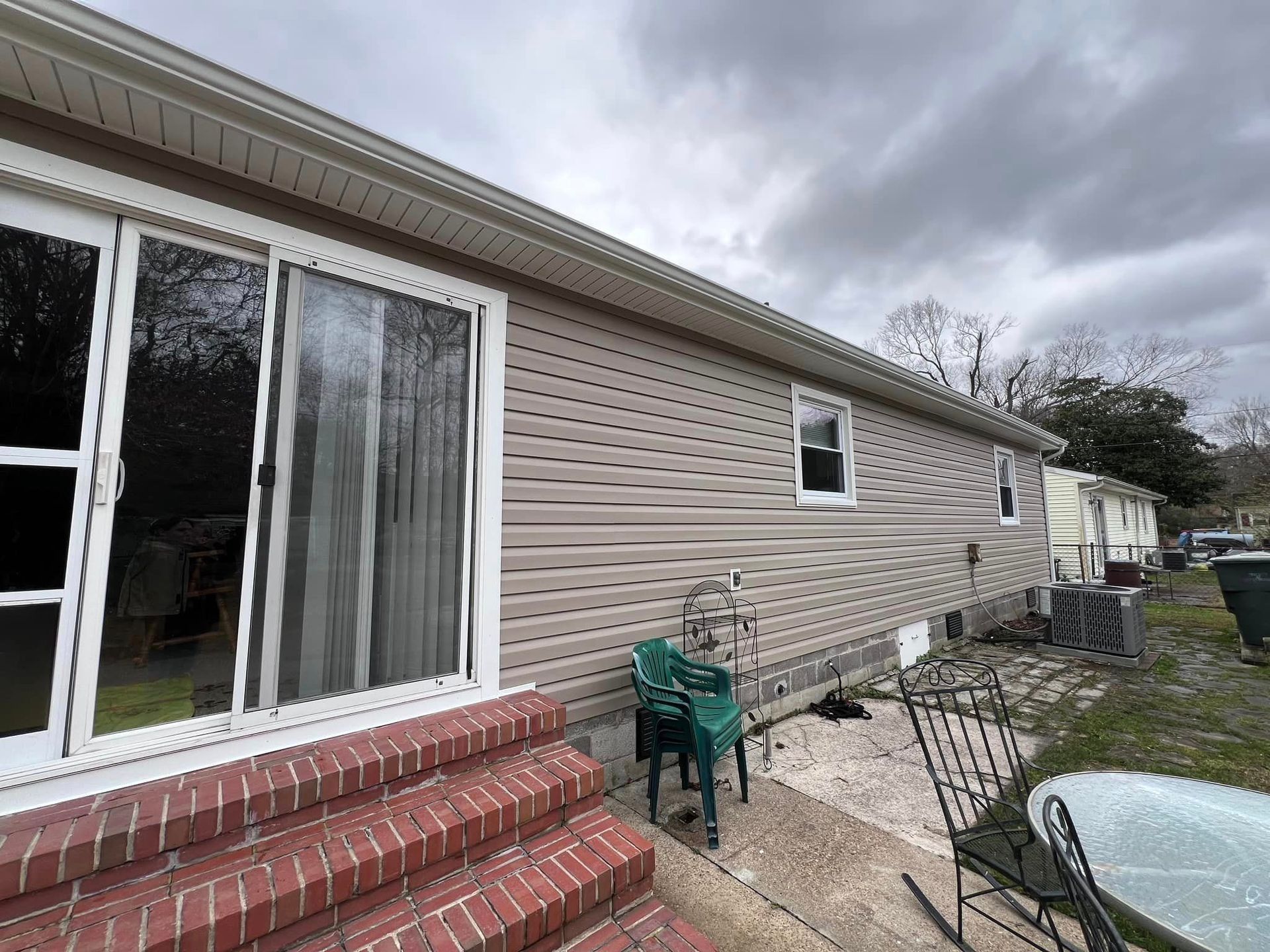Exterior view of a house with beige siding, a sliding glass door, and red brick steps. Cloudy sky overhead.