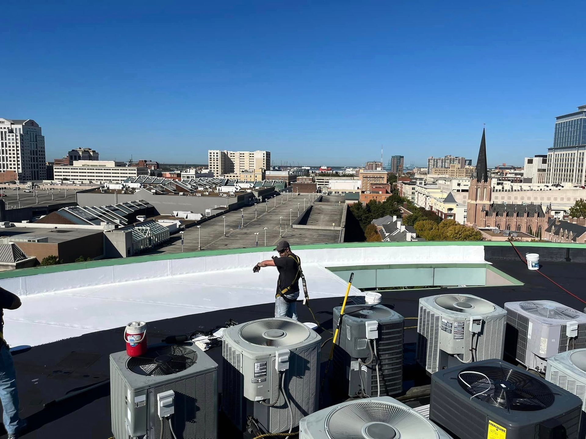 Workers on a rooftop install a white membrane. City buildings are visible in the background under a blue sky.