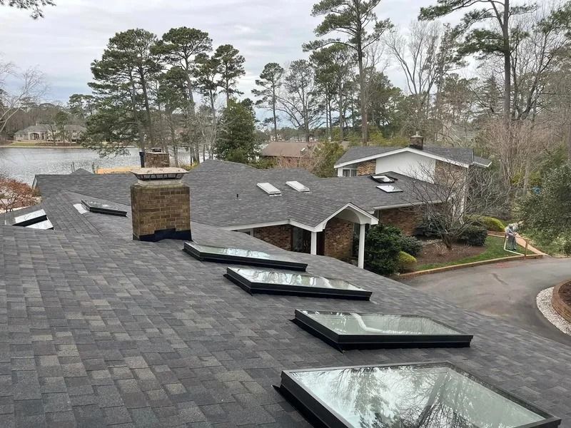 Dark gray shingle roof with six skylights and two chimneys in front of a house, trees, and water.