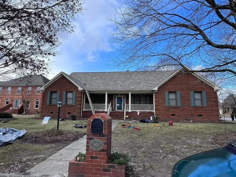 Brick house with porch, ladder on roof, mailbox, cloudy sky.