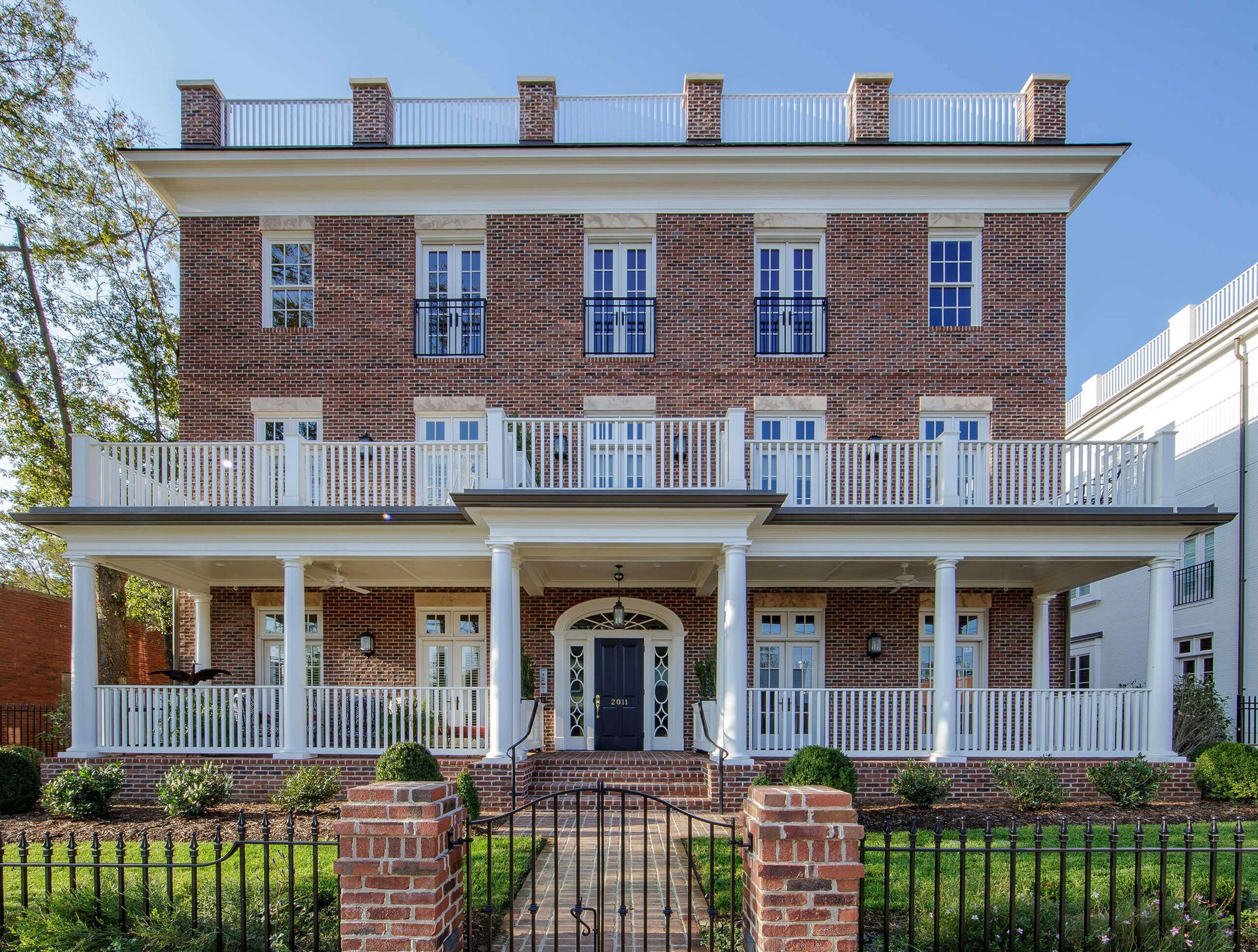 Red brick three-story house with white trim, balconies, and a wraparound porch; black iron fence in front.
