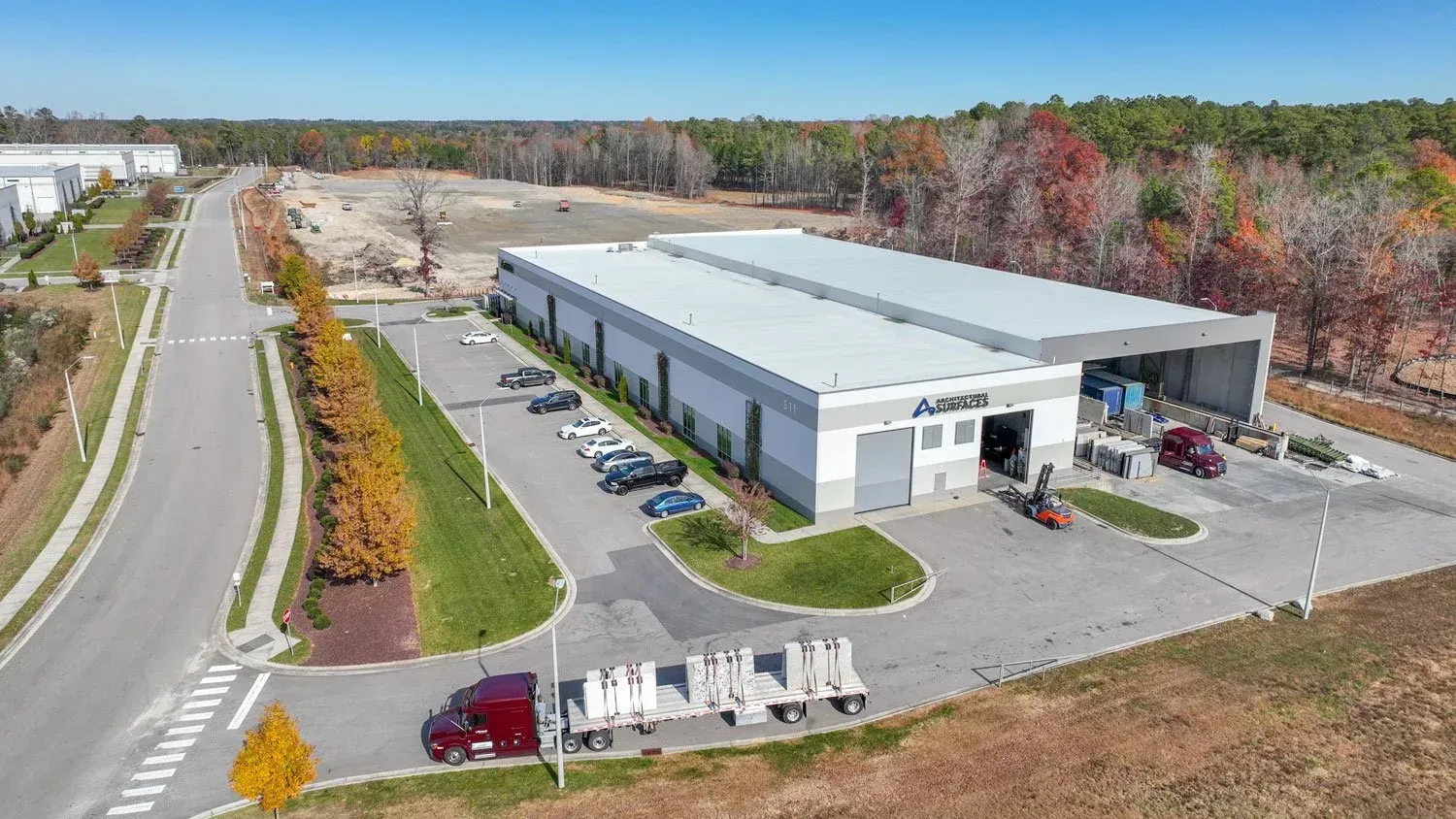 Aerial view of a gray industrial building with parked vehicles, a semi-truck, and a small forested area.