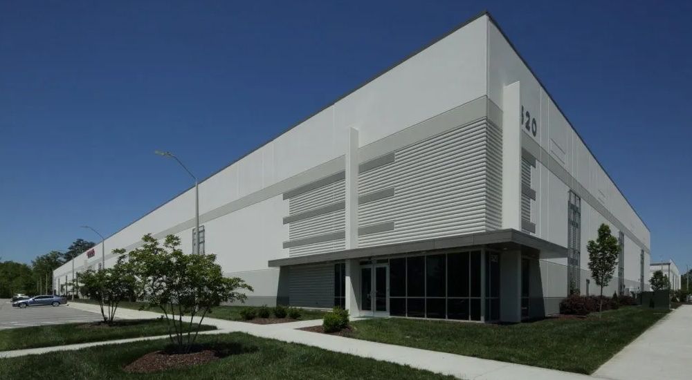 Large, modern warehouse building with white exterior, blue sky background. Green lawn and walkway in front.