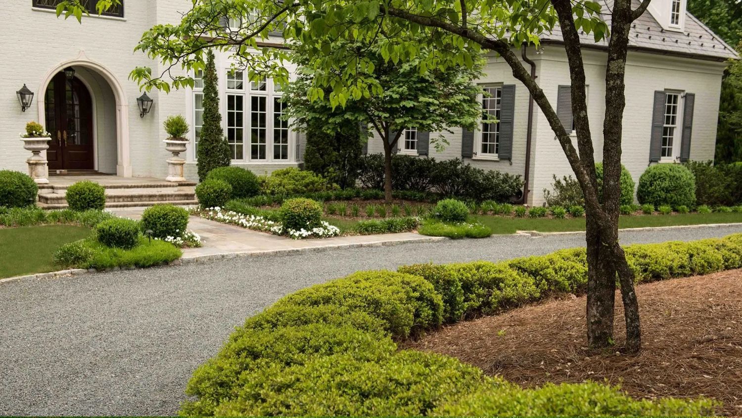 White house with arched entry, gravel driveway, and landscaped front yard with trees and shrubs