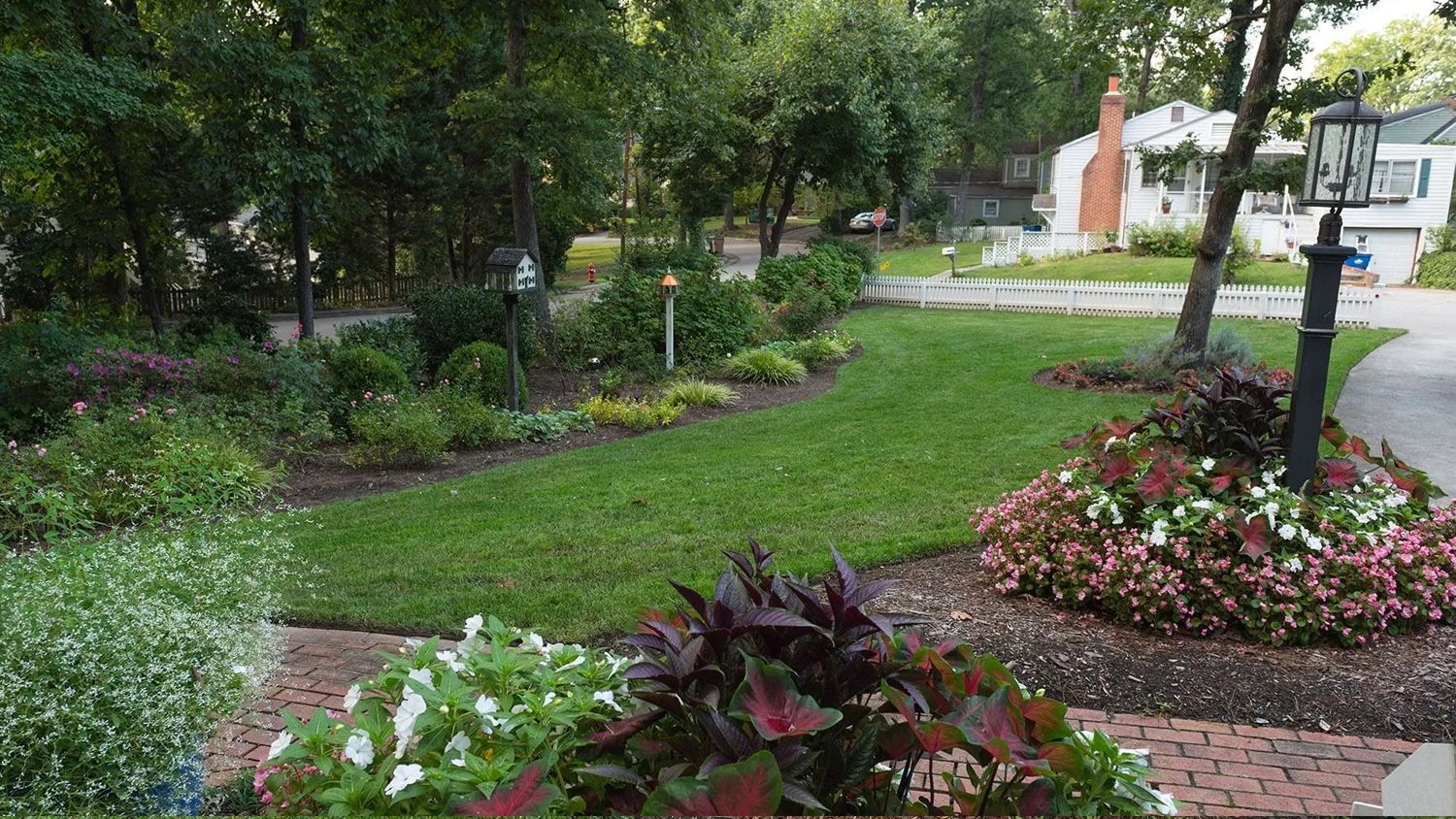 Garden path bordered by flower beds, shrubs, and trees, with a lawn and house in the background