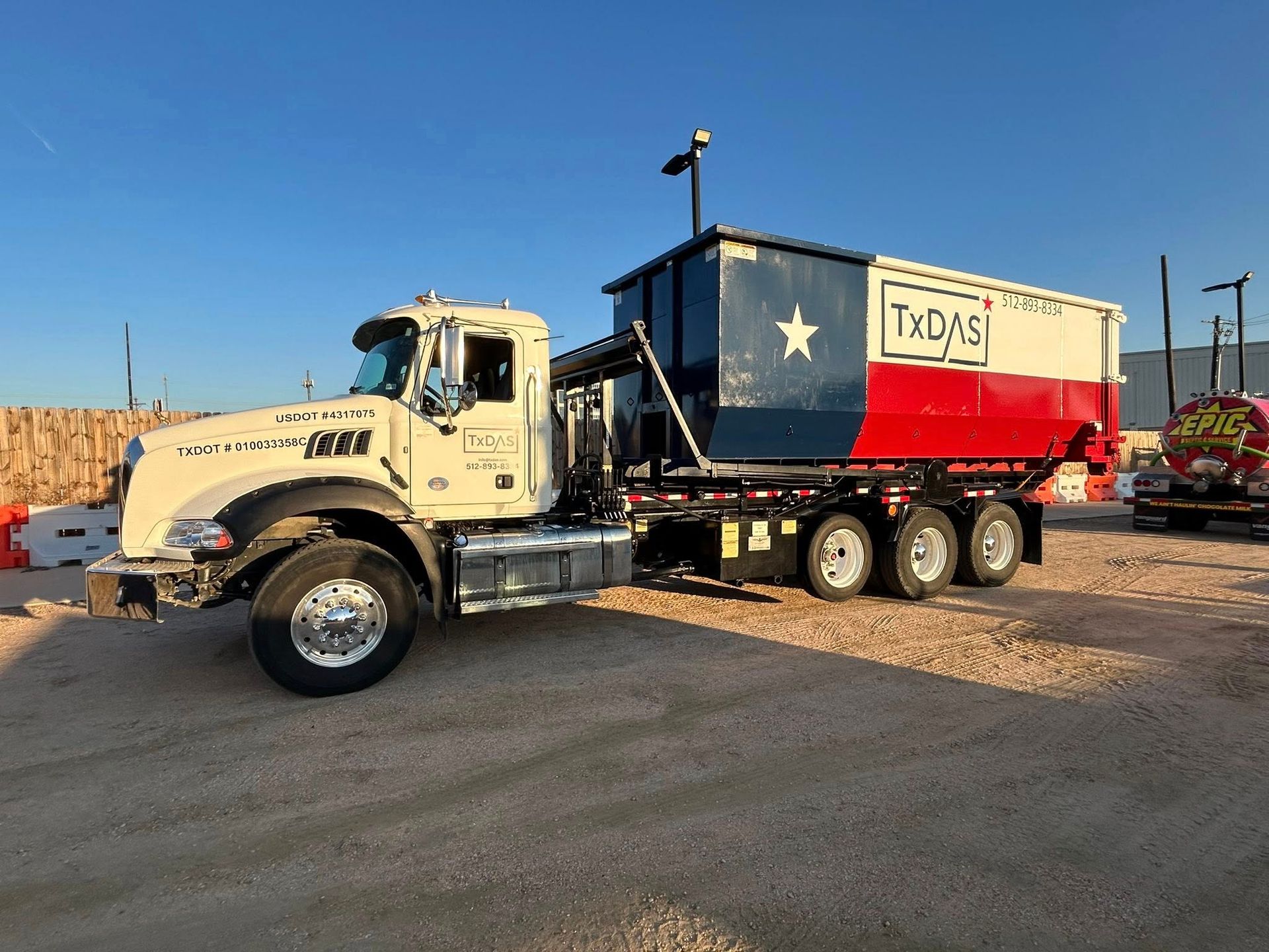 White dump truck hauling a blue, white, and red dumpster painted with the Texas flag in an outdoor setting.