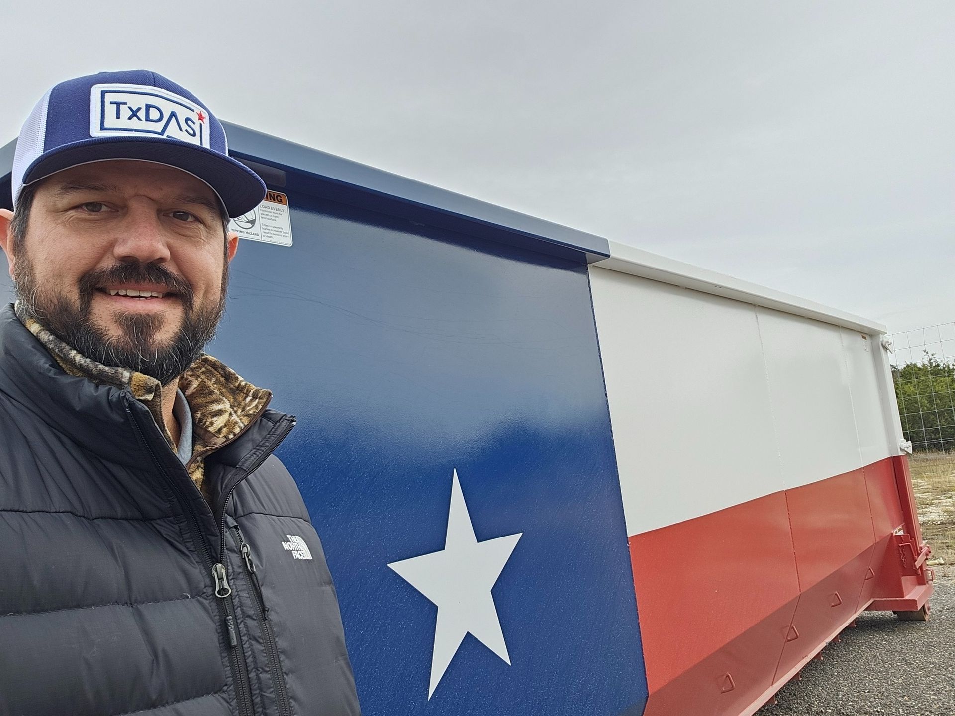 Man smiles beside a trailer painted with the Texas flag; he wears a hat that says