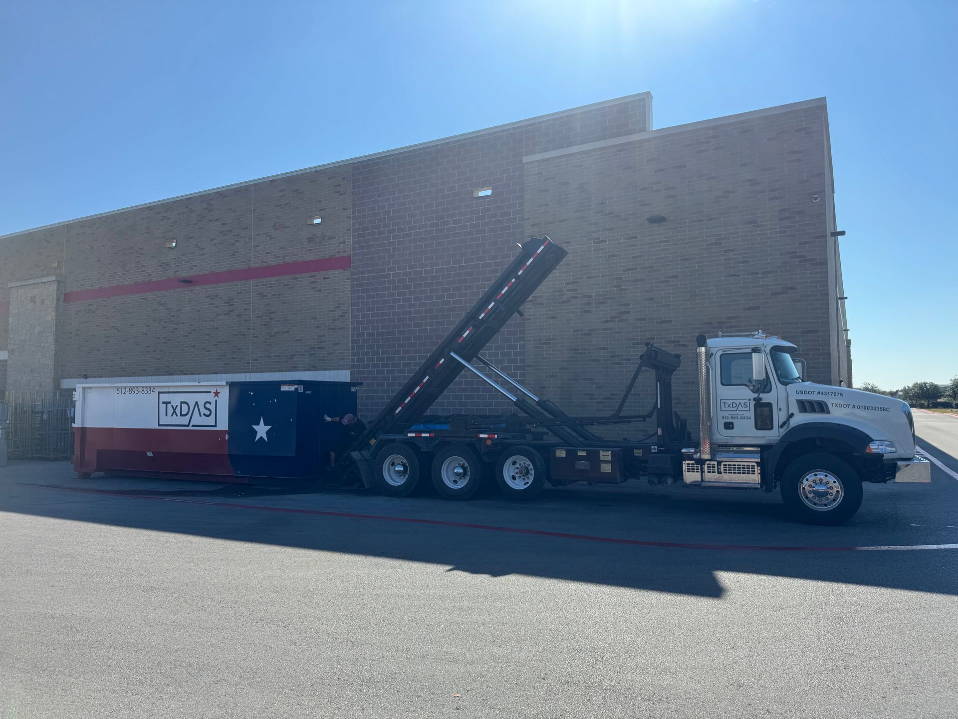 Dumpster truck with a Texas flag-themed container, parked next to a brick building on a sunny day.