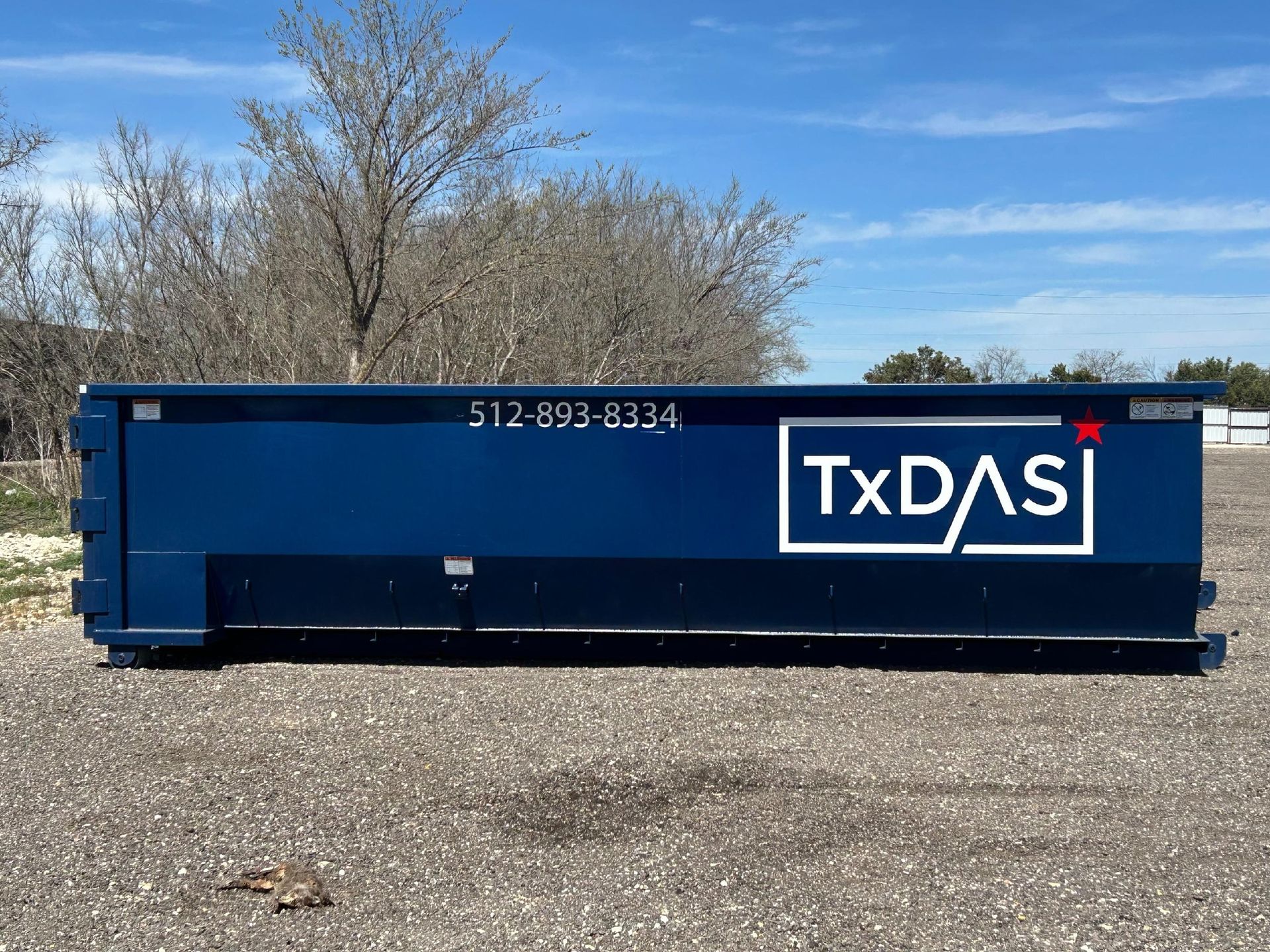 Blue TxDAS dumpster on gravel under a blue sky. Phone number 512-893-8234.
