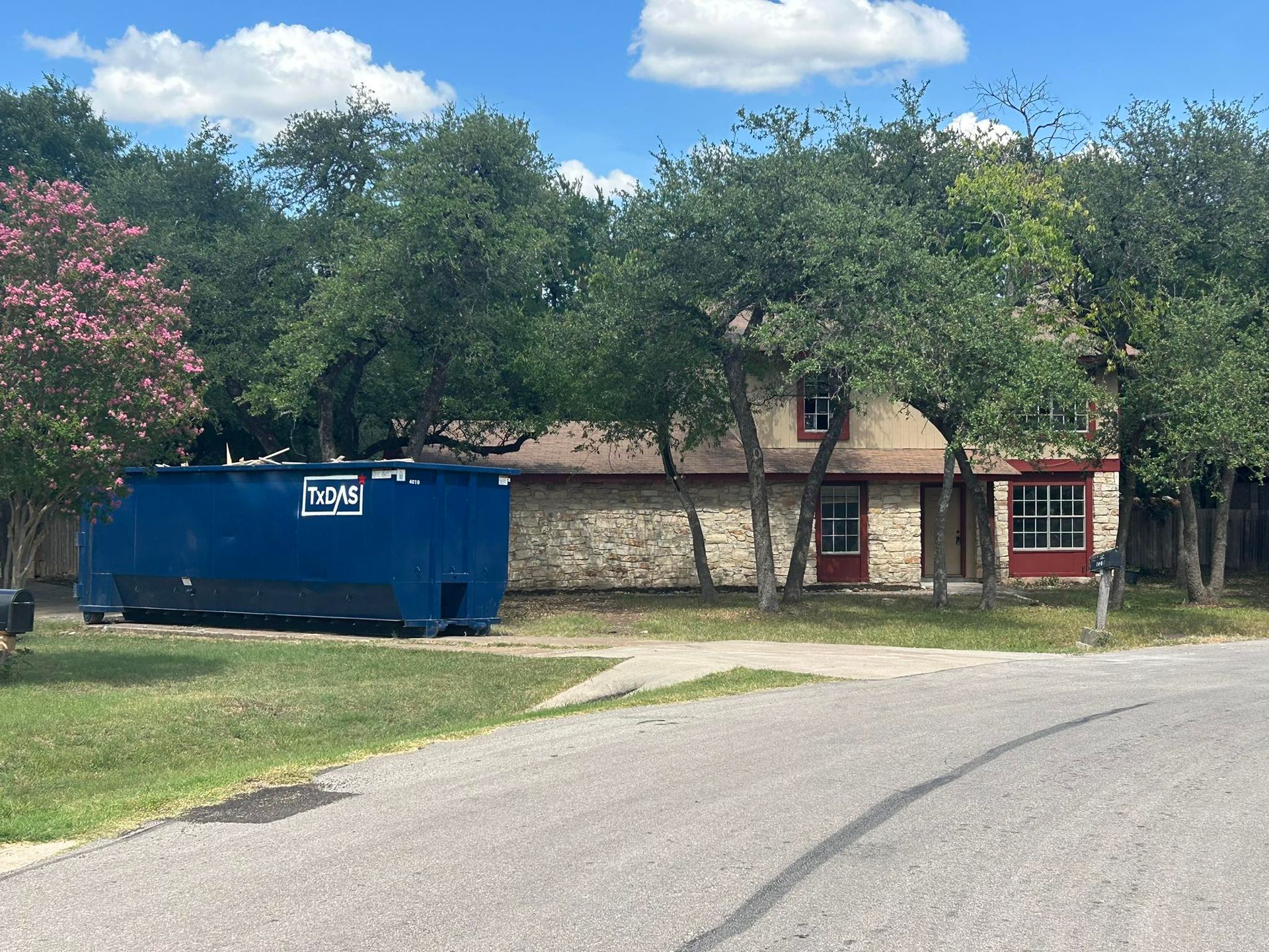 Blue dumpster in front of a two-story stone house obscured by trees on a sunny day.