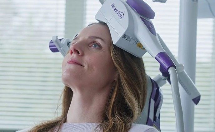 Woman using a purple-and-white facial treatment device while reclining in a bright room