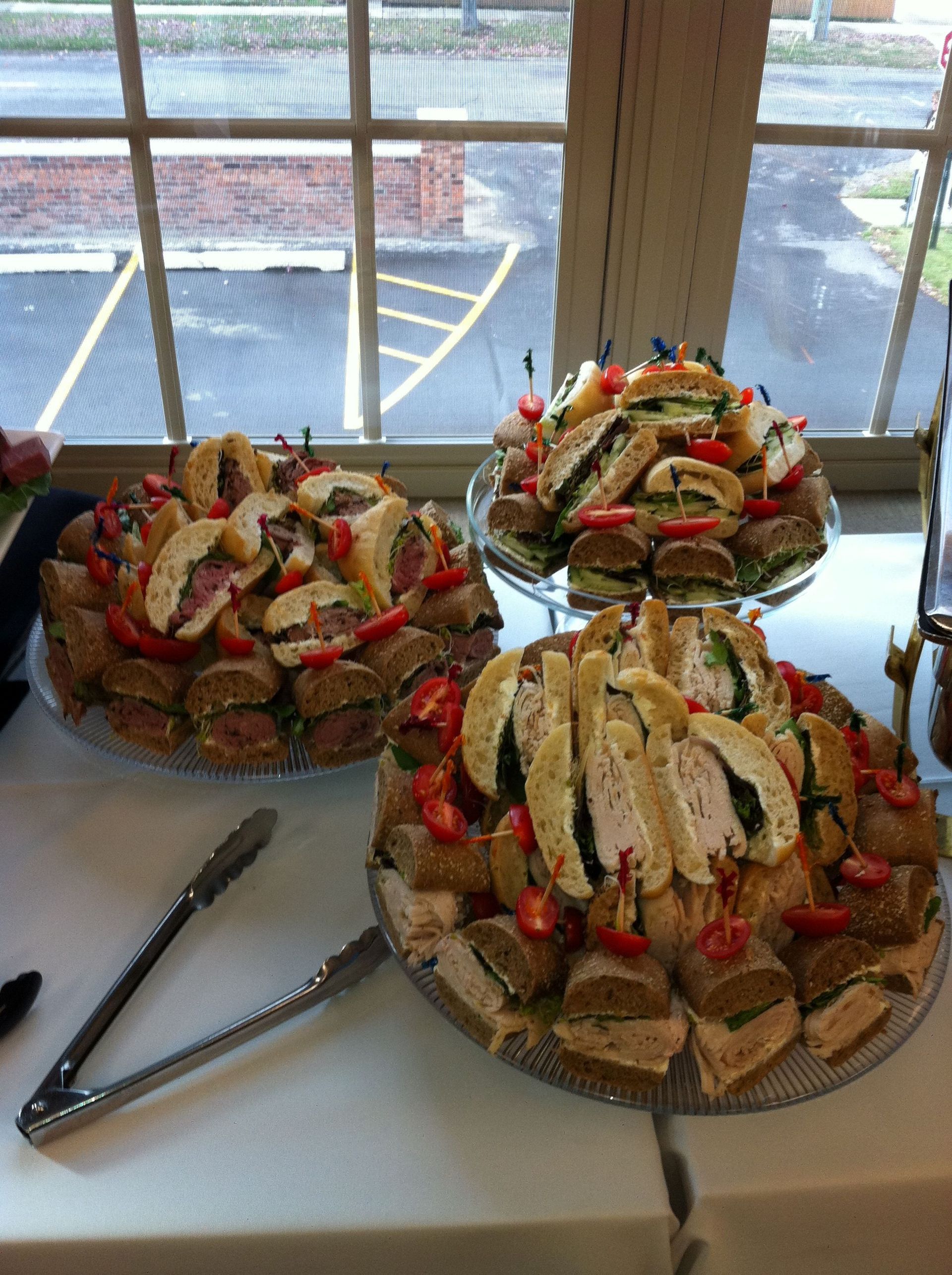 Three platters of sandwich triangles with toothpicks, set on a white table near a window.