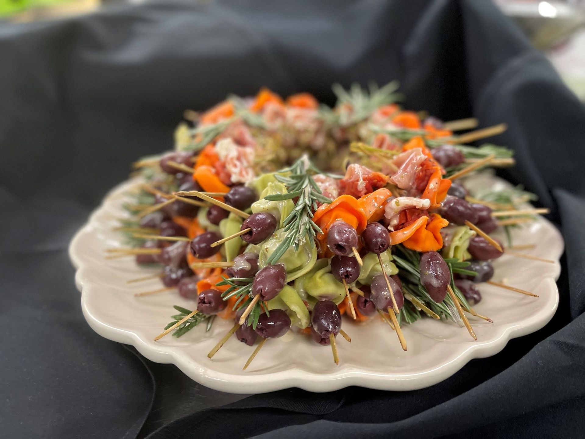Appetizer skewers with olives, carrots, prosciutto, and rosemary arranged in a circular wreath on a scalloped plate.