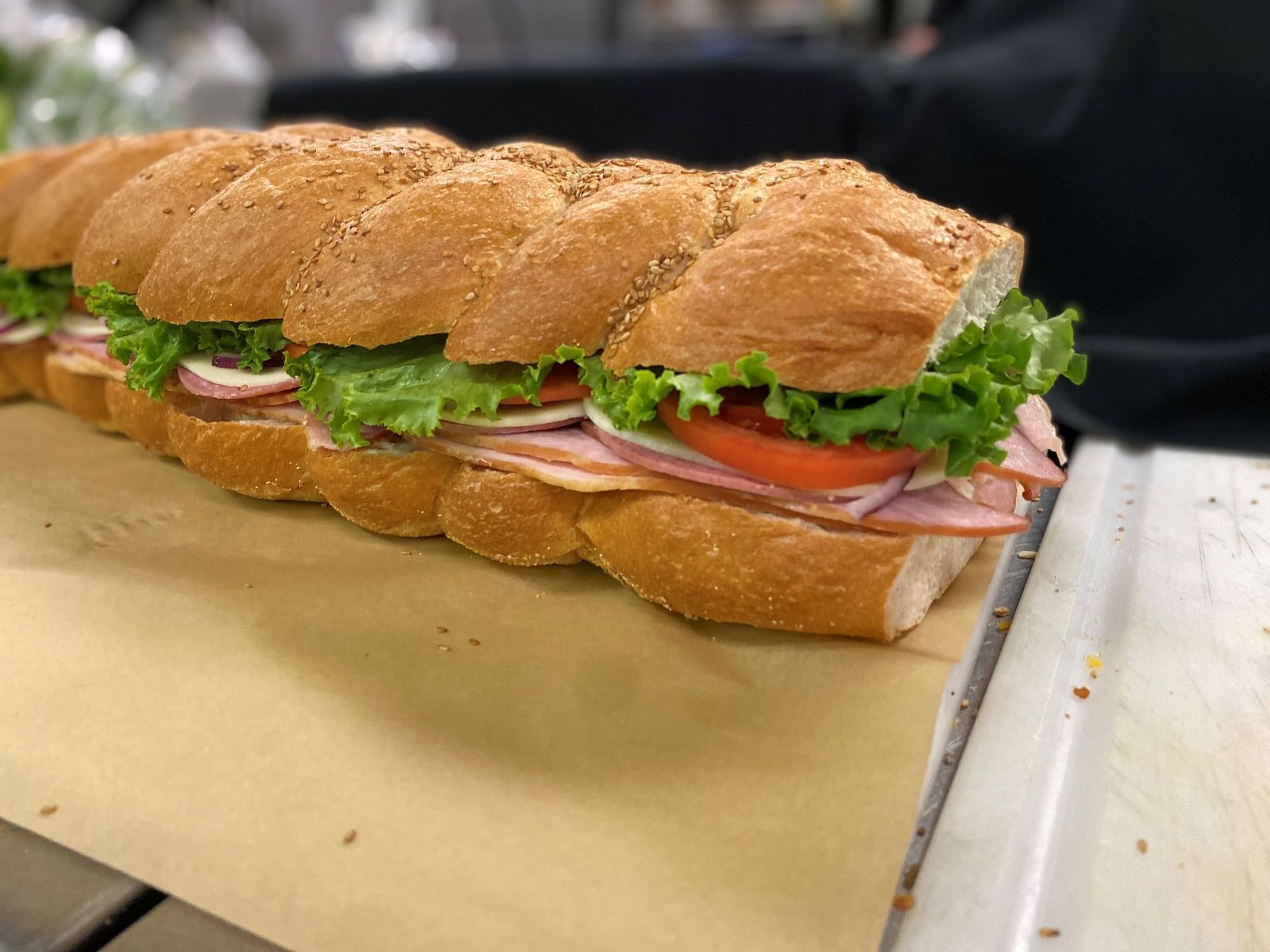 Long sandwich with ham, tomato, lettuce, and onion on a brown loaf, on a counter.