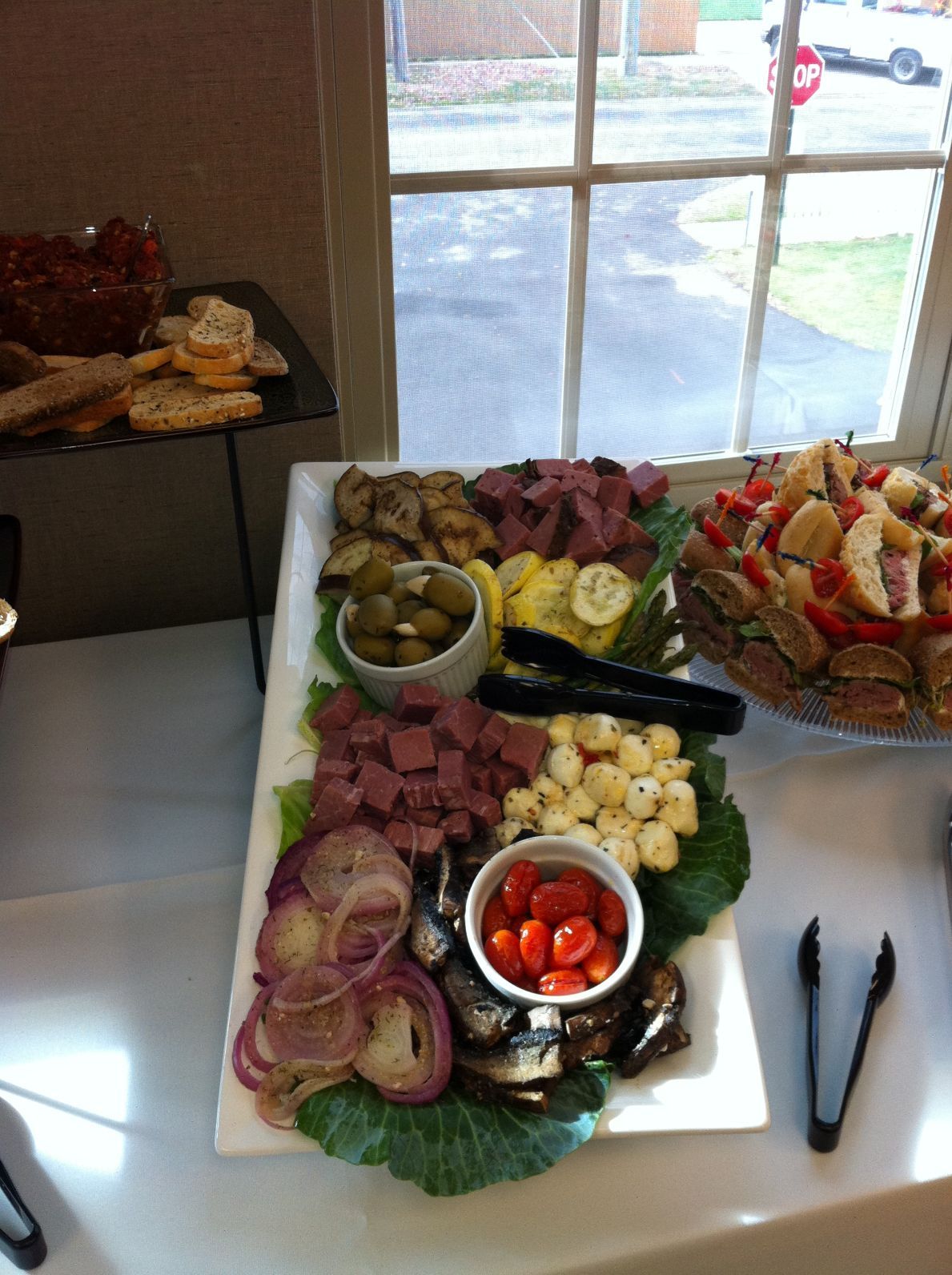 A platter of food with sliced meat, olives, tomatoes, and pickled onions on a white table near a window.