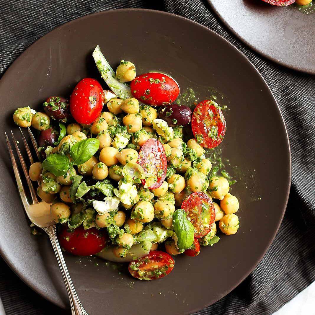 Chickpea salad with tomatoes, olives, and pesto on a brown plate, fork on the side.
