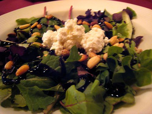 Salad with greens, goat cheese, pine nuts, and balsamic glaze on a white plate.