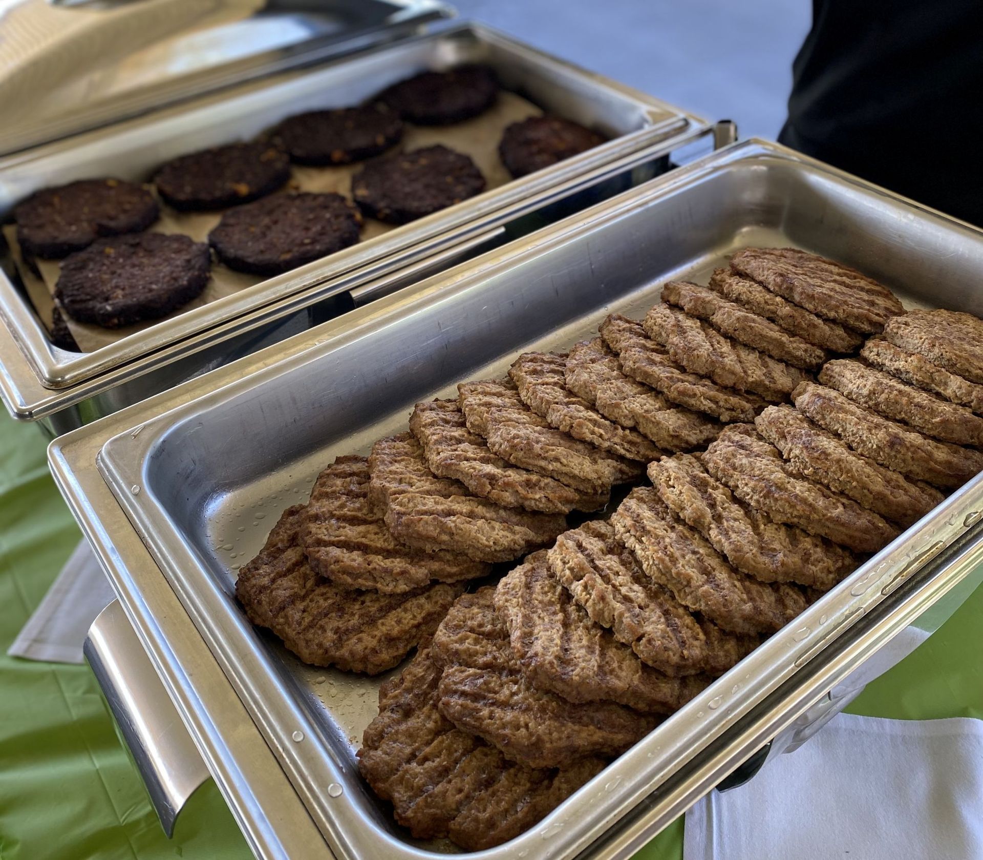 Two stainless steel food trays, filled with cooked hamburger patties.