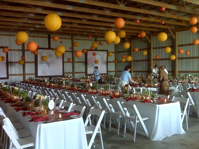 Barn interior set up for a celebration: tables with white linens, red accents, orange lanterns. People setting up.