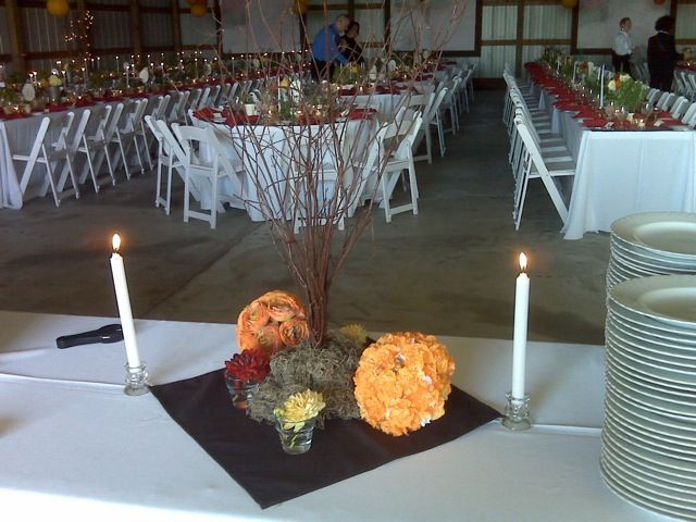 Long tables set for a celebration in a barn; centerpieces include candles, pumpkins, and branches.