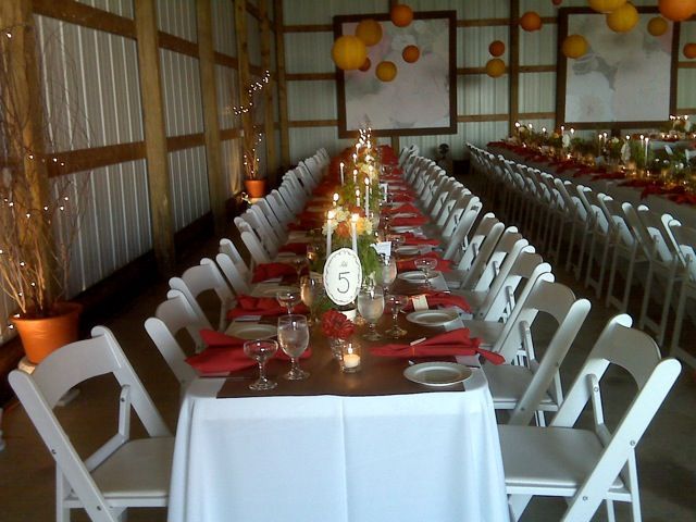 Long table set for a wedding reception in a barn, white chairs, red napkins, candles, and orange paper lanterns.