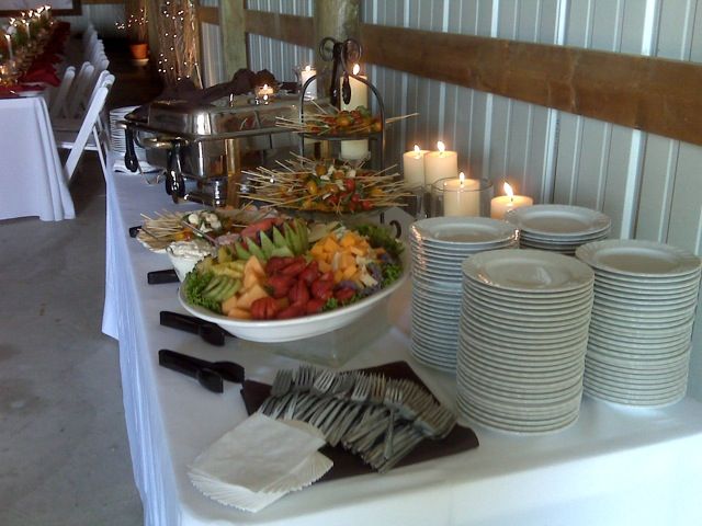 Buffet table with food, candles, and stacks of plates in a room with white walls and a metal roof.