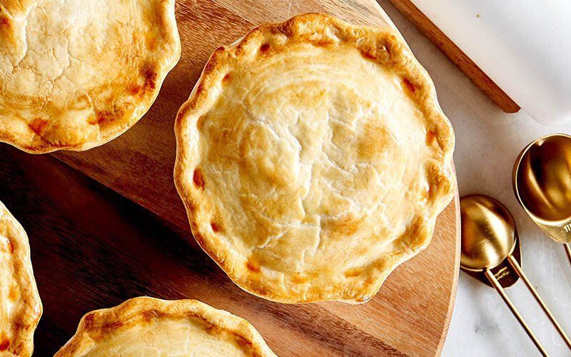 Golden-brown, round pot pies with crimped crusts on a wooden board; measuring spoons nearby.