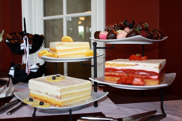 Dessert display: three-tiered stand holding layered cakes, chocolate-covered strawberries, and muffins, near a window.