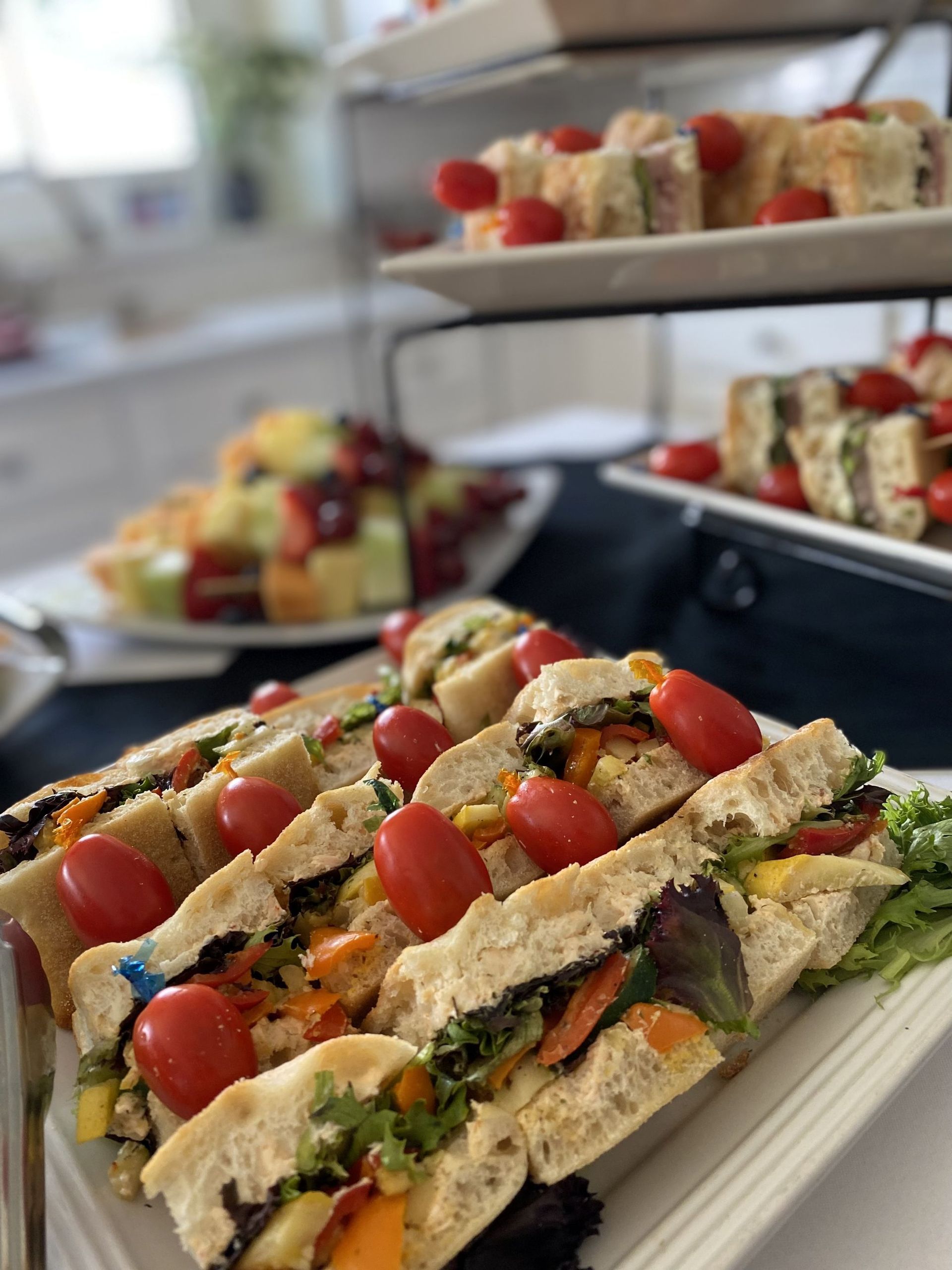 Sandwiches on tiered serving platters with cherry tomatoes, fruit platter in background.