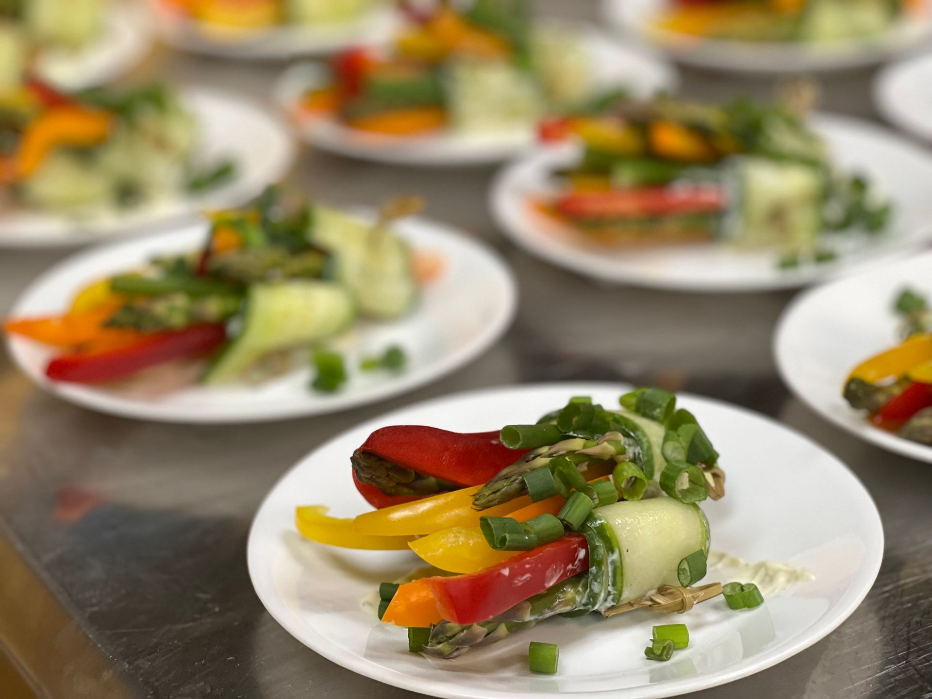 Many small white plates on a metal counter, each holding a fresh cucumber-wrapped vegetable bundle topped with scallions.