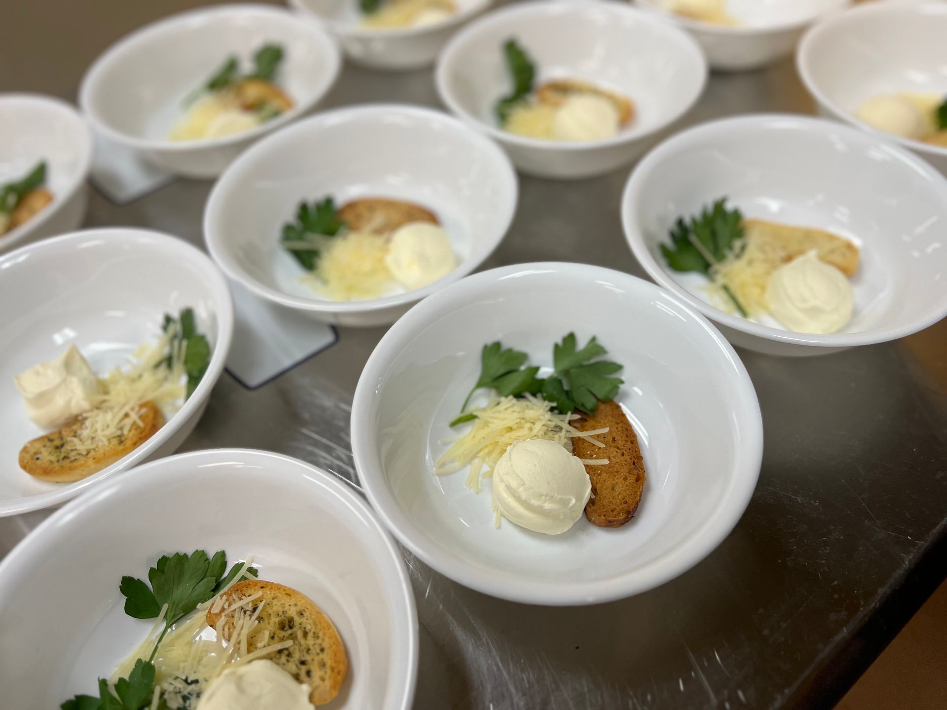 Several white bowls arranged on a surface, each containing a crostini, a dollop of cream, shredded cheese, and parsley.